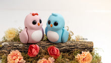 Two small bird figurines, one pink and one blue, sitting on a log with flowers around them.