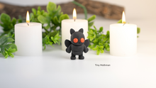 Black creature figurine between three lit candles with greenery in the background